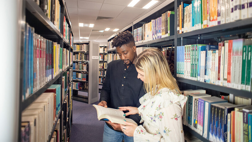 Two students browsing through a book in The Sir Michael Cobham Library. The Library is home to thousands of printed collections, alongside a large collection of e-books and e-journals