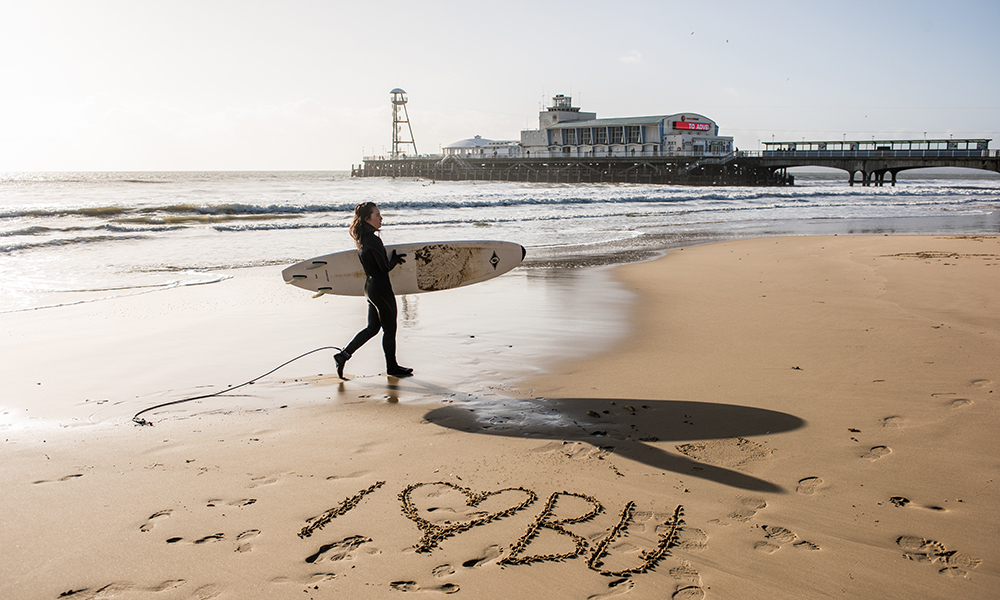 Woman in a wetsuit carrying a surfboard walking across the beach with 'I heart BU' written in the sand