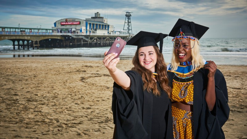 Two female graduates in gowns and caps on the beach with the pier and sea in the background