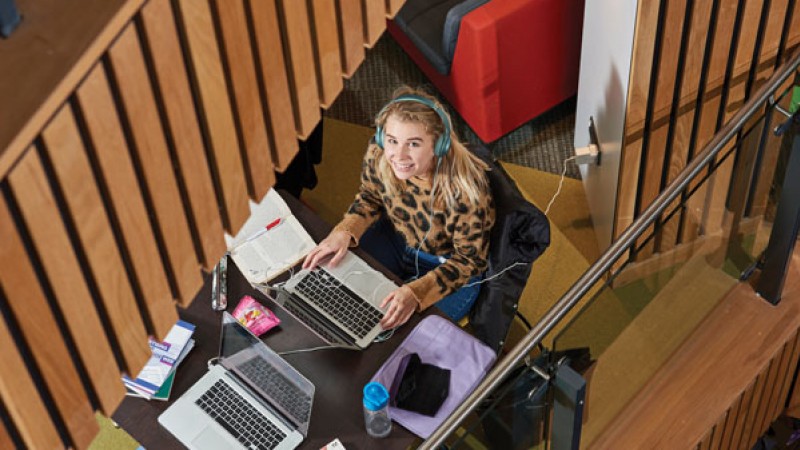 A student in the Student Centre, working at a laptop and looking up at the camera, smiling