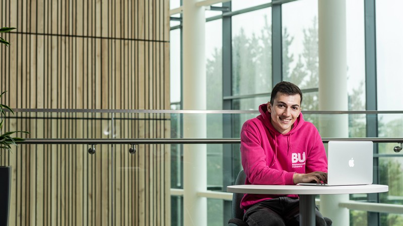 A student ambassador with a laptop on a table, smiling at the camera in the Bournemouth Gateway Building