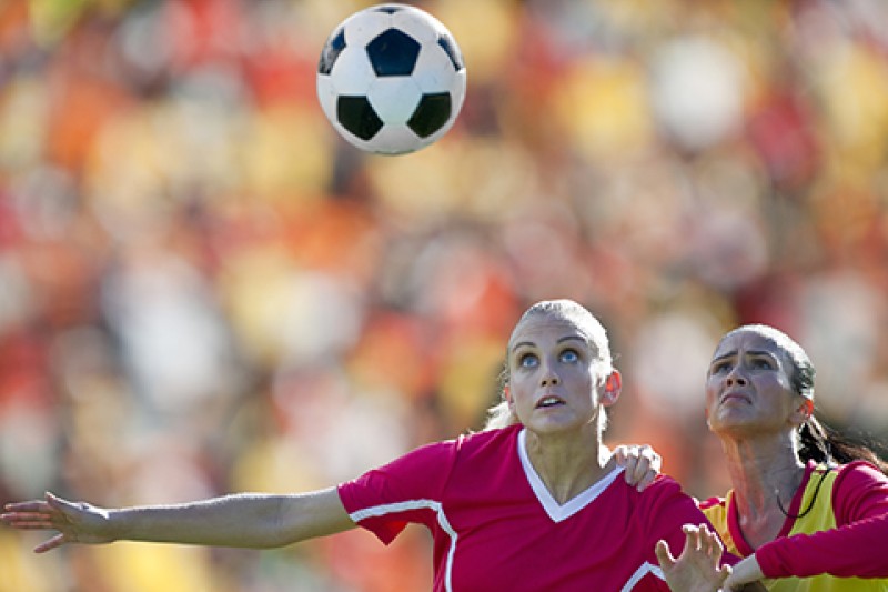Two women playing football on opposite teams, looking at the ball which is in the air above their heads
