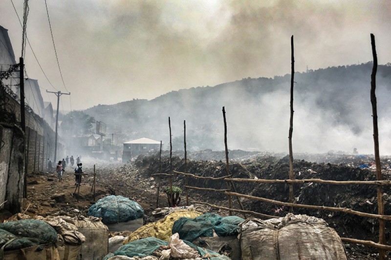 Bags of rubbish on a landfill site in Freetown, Sierra Leone, with smoke rising from piles of rubbish in the background