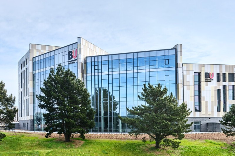 Bournemouth Gateway Building is lit by the sun on a clear day. the grass verge and trees are shown infornt of the building with the BU logos large and bright on two corners of the external structure.