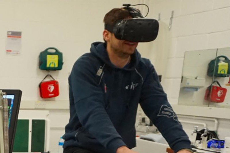 Creative Technology research ongoing in a lab with a man on an exercise bike, wearing a VR headset, with a woman observing activity on a monitor