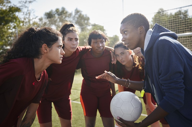 A female coach, holding a football, in a huddle talking to four female players
