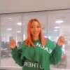 Esther poses with her arms in the air, wearing a green jumper and black leggings. the backdrop is a room on campus