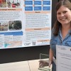 Malika holding her certificate and trophy in front of a poster depicting her research