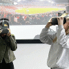 Two Bournemouth University students wearing virtual reality headsets, with a screen showing a football game in the background.