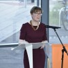 Professor Katharine Cox stands infront of a lectern and microphone, wearing a read dress and statement necklace. She addresses an audience to introduce guest speakers. 