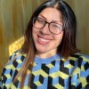 Zoey smiles to the camera in a selfie style photo, the light is on her face as she smiles. Zoey wears her glasses and a checked jumper in black, light blue, and yellow 