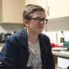 student Jonathan is pictured in a kitchen, wearing a navy blue jacket and a white t-shirt. he wears glasses and a smile on his face