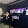 Four students sit in front of a wall of TV screens, each showing multiple broadcasts