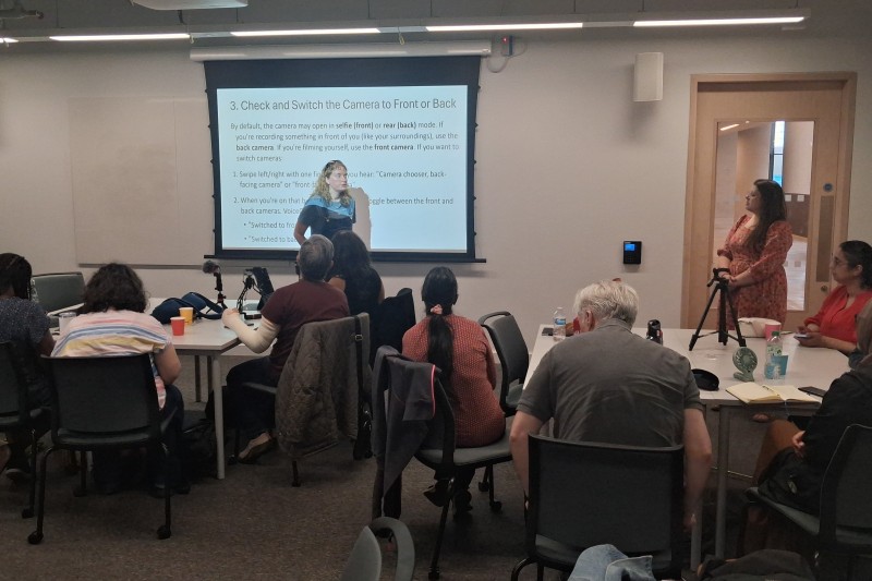 Blindness and media creation research project workshop in action at Bournemouth University. People are sat on tables in a seminar room with a projector screen and a project participant is leading the workshop