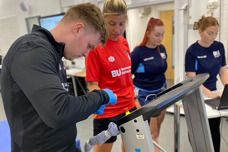 An AFCB Women's team player slowly walks on a treadmill whilst 3 members of the Bournemouth University team look at a computer screen linked to the treadmill