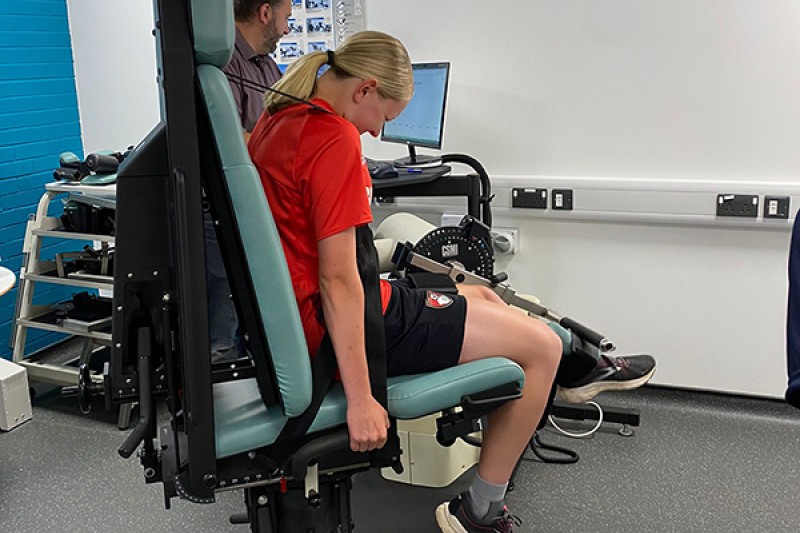 Football player sits in a chair lifting a weight with her left leg whilst a trainer monitors a screen linked to the fitness equipment