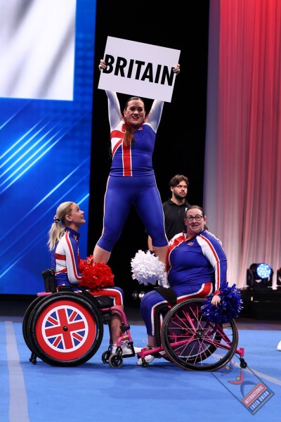 Abbie and another team member, both in wheelchairs, supporting a colleague who is standing and holding a card with "Britain" on it
