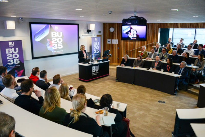 Alison standing at a lectern talking to a room of guests with the BU2035 logo behind her. 