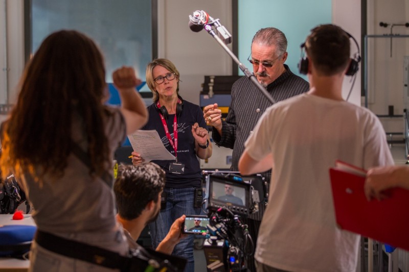 Griff Rhys-Jones stands in from of a camera. To his left is the director assistant. In front is the film camera with griff's image on the small display. A boom operator stands at the front with his back to the camera holding up the boom mic