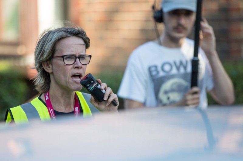 A lady wearing a high-viz tabbard speaking into a radio. Behind her, a boom operator holds the boom mic