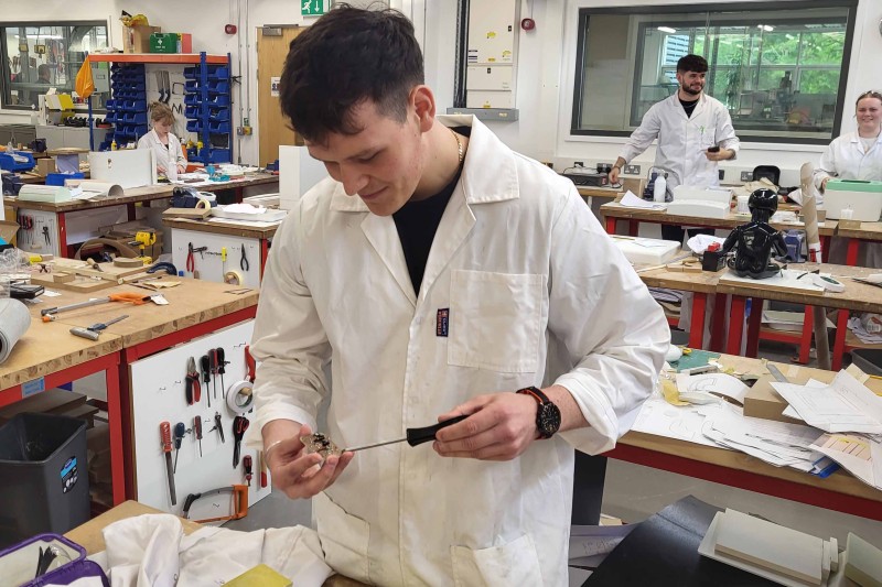 Archie in the workshop with an electrical component he is working from in one hand and a tool in the other