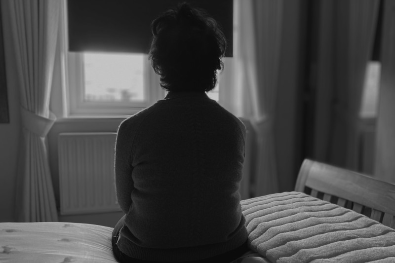 A photograph of the back of a lady sitting on the edge of her bed looking out of her window