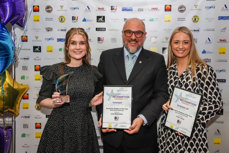 A male and two females looking at the camera smiling. The man in the middle is holding a framed certificate. The woman on the left of the picture is holding a trophy and the woman on the right of the photo is also holding a certificate