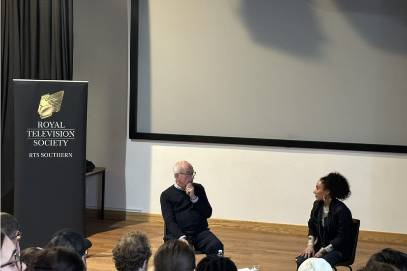 Yasemin Rashit A man and woman sitting on chairs speaking to each other, students sit in the seats in front of the stage