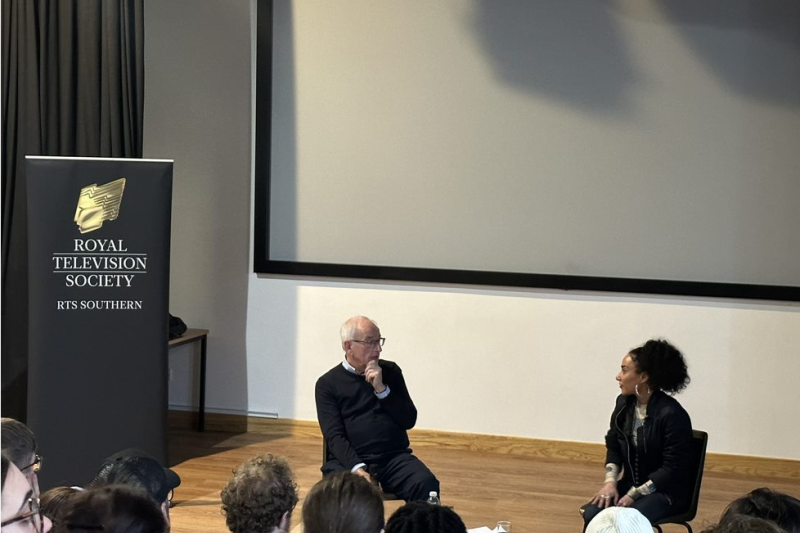 A man and woman sitting on chairs speaking to each other, students sit in the seats in front of the stage