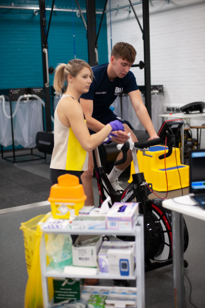 A man pedals on an exercise bike, Becky Neal stands next to him looking at a screen connected to the bike