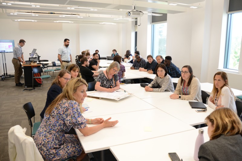 Seven women sitting around a table having a group discussion. One person is making notes on a flip chart. Other groups of people sit around tables in the background