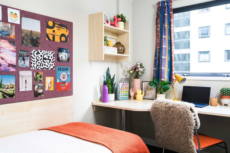 The bottom half of a single bed with a pin board on the left wall and a desk facing a window
