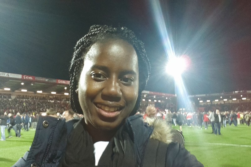 A selfie of Brigitte on a football pitch with ground on the pitch behind her and a floodlight shining in the background