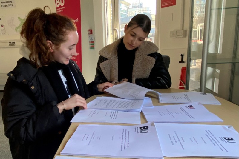 Two female students sitting at a table looking through piles of paper