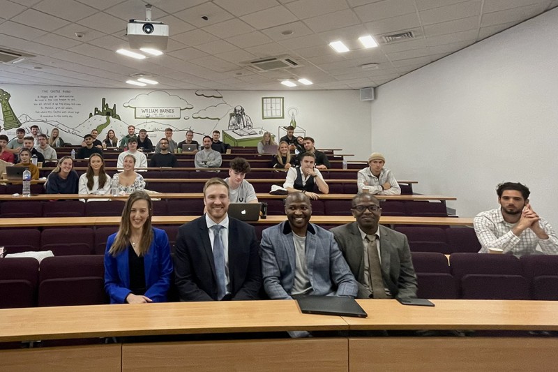 The four panellists sitting in the front row of a lecture theatre smiling at the camera. Students sit on the rows behind them.