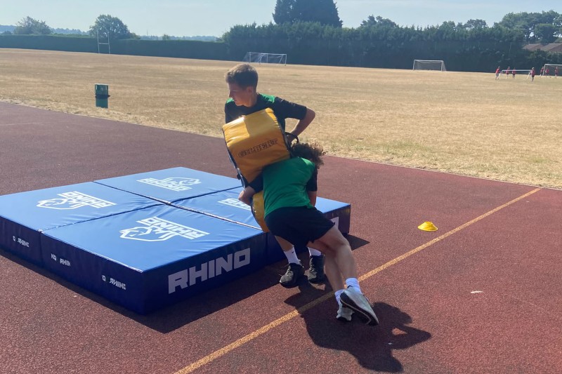 A young boy charging into a rugby tackle pad being held by his teacher