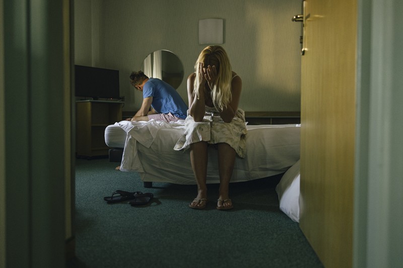 Couple A woman sits on the side of a bed facing the camera, covering her face with her hands. A man sits on the other side of the bed, looking away from her