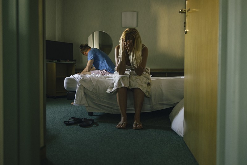 A woman sits on the side of a bed facing the camera, covering her face with her hands. A man sits on the other side of the bed, looking away from her