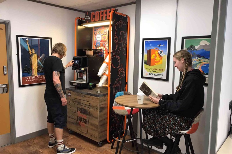 Two students are relaxing in the library's coffee cabin. One is choosing a drink, and the other is seated reading a book