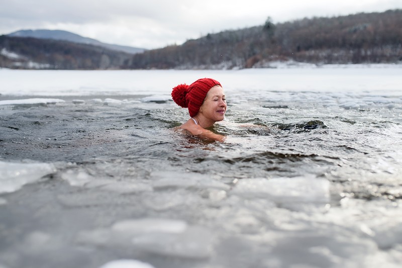 A lady swimming in an icy lake wearing a woolly hat, hills are in the background