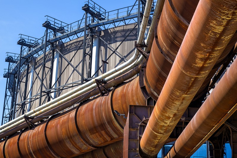 Some large, rusty pipelines running across the photo with a large gas holder in the background