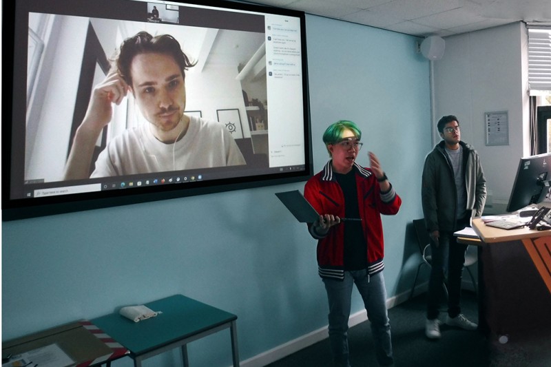 A man standing in front of a class room, addressing students, holding his laptop