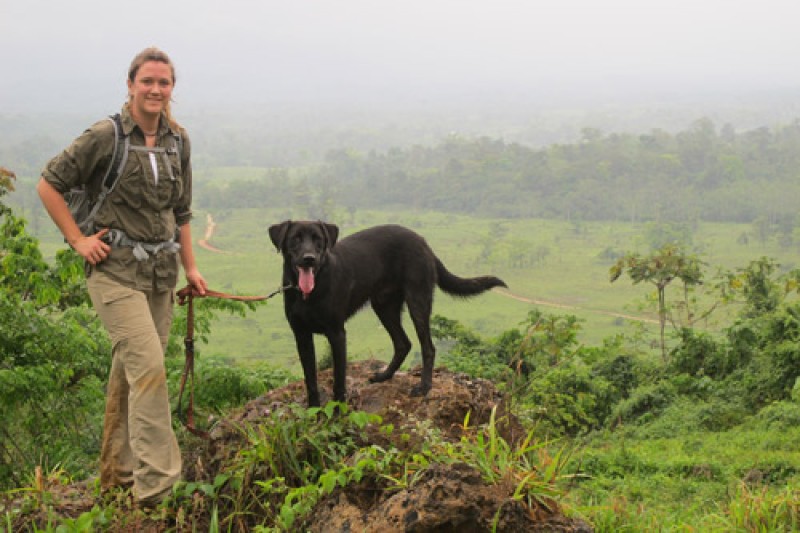 Dr Jennifer White with a dog on a lead standing on a high vantage point. Open green fields and trees in the background