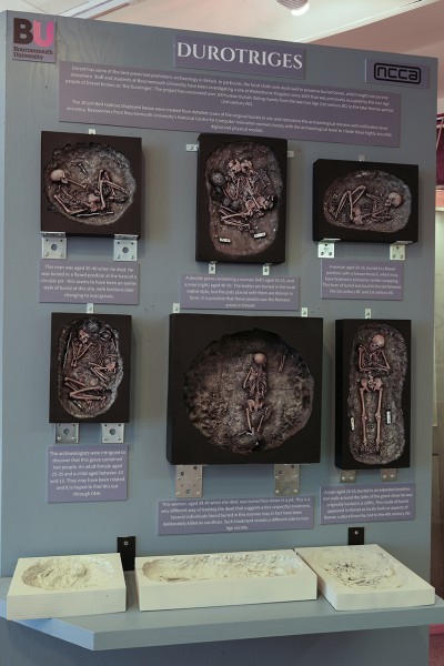 A photo of the display in the museum. 6 3D models showing ancient skeletons in their graves have been mounted on to the wall. In front of the exhibit is a shelf holding 3 more 3D printed models which can be picked up and handled. 
