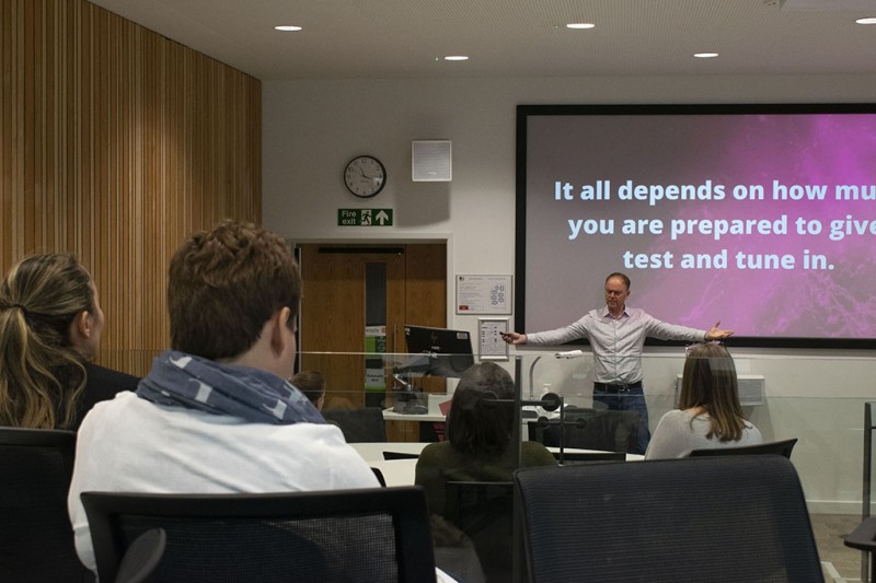 A man at the back of the room facing towards the camera talking in front of a projector screen. The backs of students sitting in the room listening to him are in the foreground