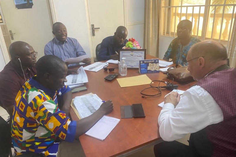 Six men sitting around a table. At the far end a man is reading out from a sheet of paper