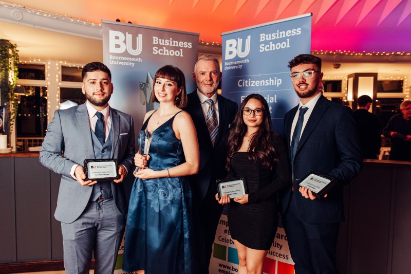 Two female and two male students in smart attire holding their awards in front of them, standing in front of a Business School banner