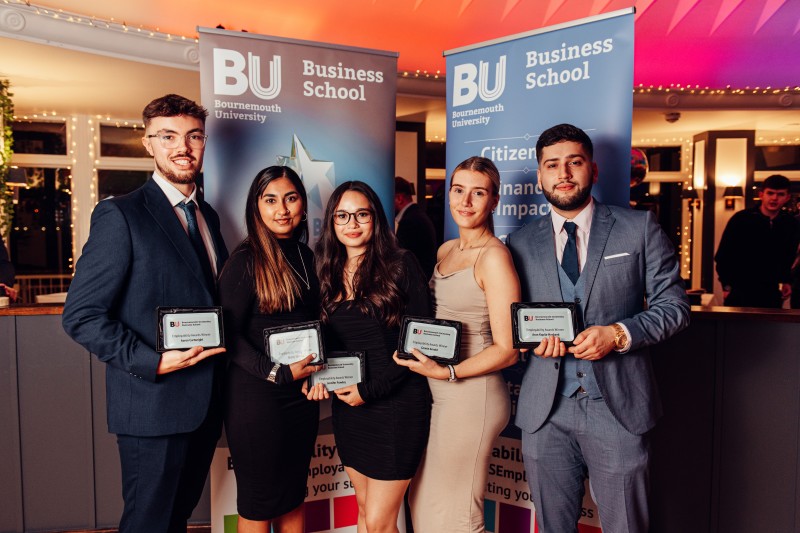 Three female and two male students in smart attire holding their awards up in front of a Business School banner