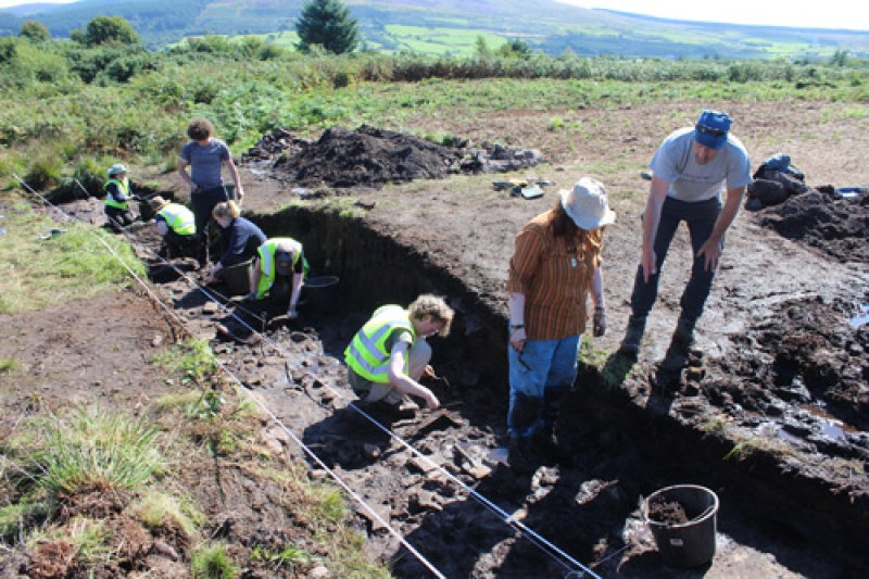 Severn people in a large trench that they are excavating in a field. Rolling hills and fields are in the background