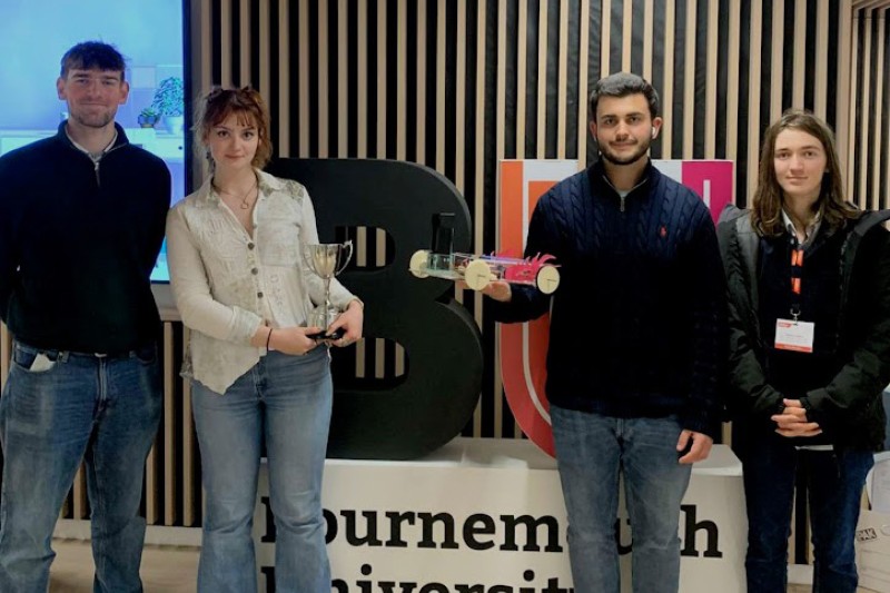 Three male and one female student standing in front of the BU logo, one holding the winning trophy and one holding their robot